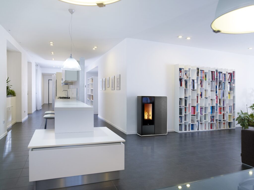 Intérieur moderne et lumineux avec cuisine blanche, îlot central, poêle à granulés noir, et grande bibliothèque modulable. Sol carrelé sombre.
