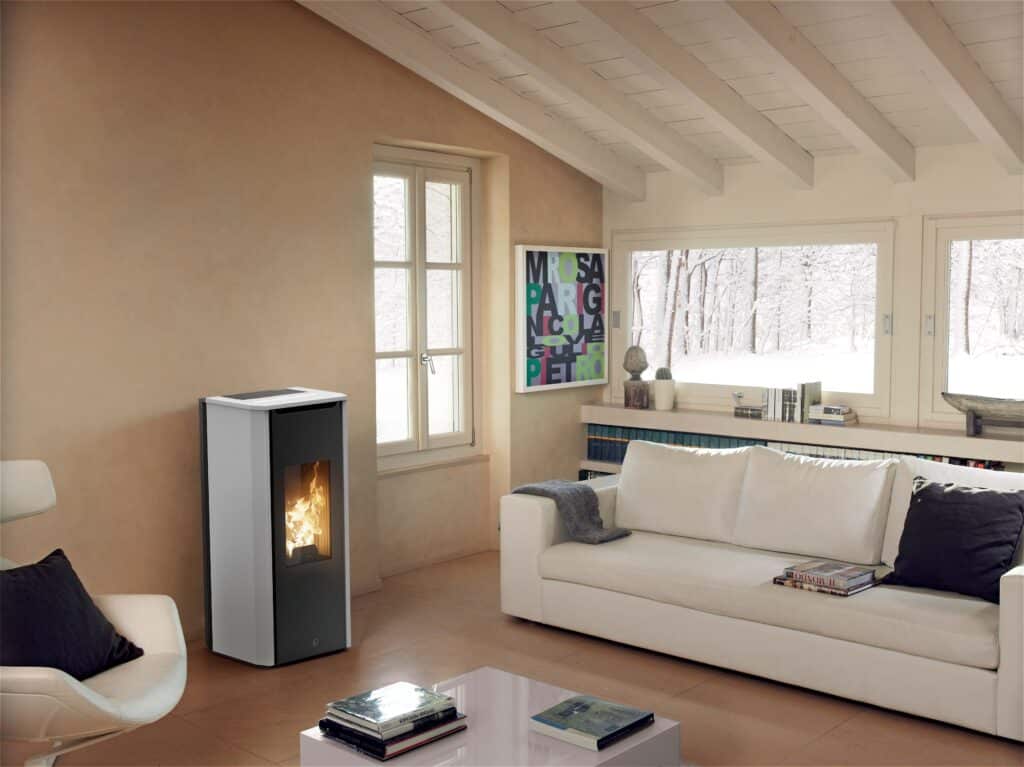 Salon cozy avec poêle à granulés moderne allumé. Canapé blanc, fauteuil, table basse. Fenêtres sur forêt enneigée, plafond poutres.