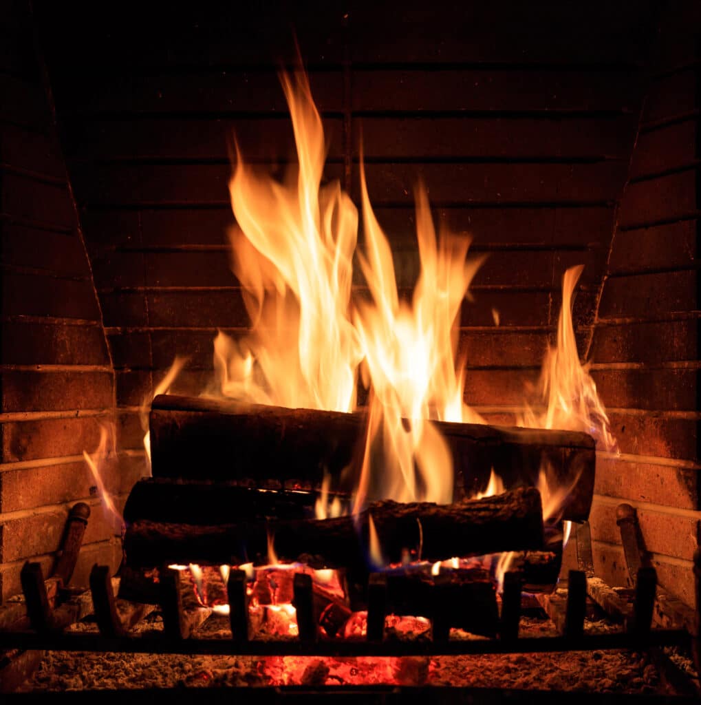 Cheminée en briques sombres avec de grandes flammes vives. Des bûches brûlent sur une grille en fer, créant une ambiance chaleureuse et lumineuse.