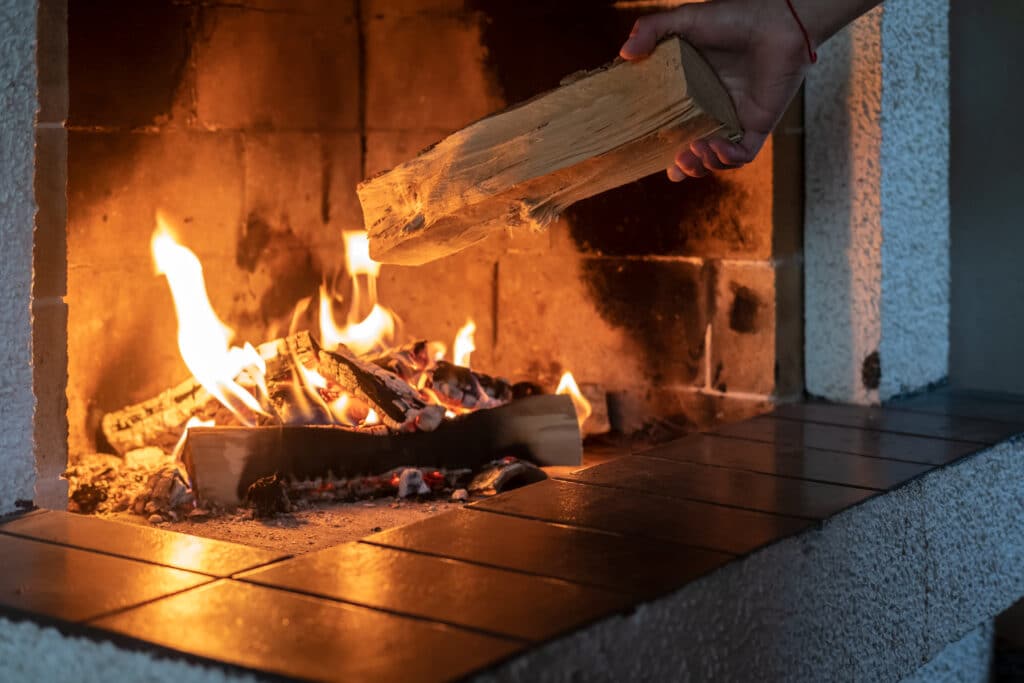 Main plaçant une bûche dans une cheminée où un feu ardent crépite. Lumière chaude des flammes sur l'âtre.