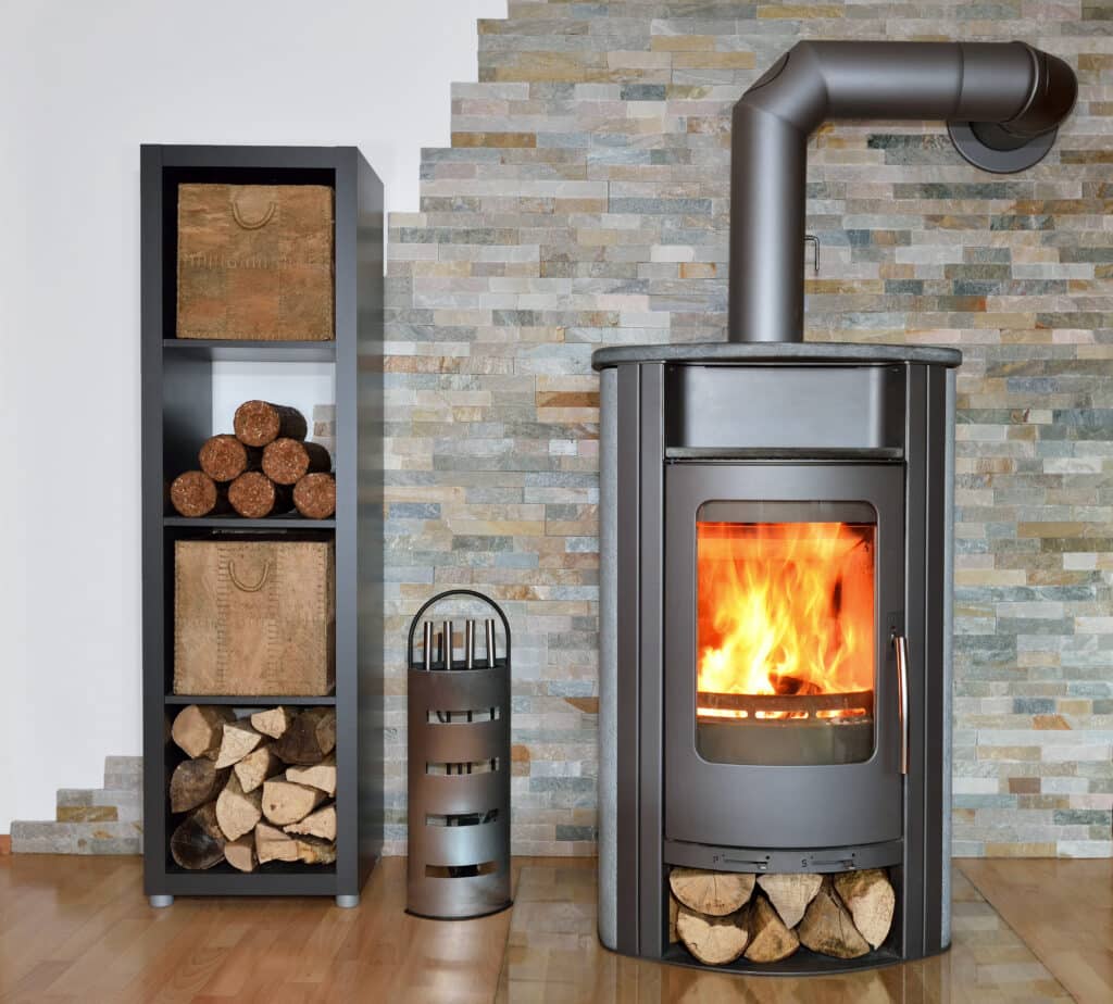 Poêle à bois moderne anthracite avec flammes vives, adossé à un mur en pierre naturelle. À gauche, une étagère sombre contient bûches et paniers. Plancher en bois.